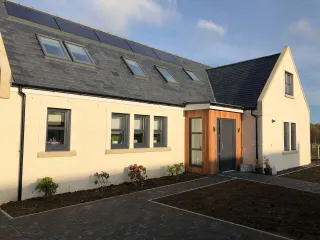 the front aspect of a classic Scottish rural cottage with cream coloured facade, a wood clad front porch with aluminium door and various aluminium windows 