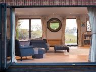 fully open bifold door from the outside looking in to a room with sofas, foot stools, wood flooring and glass windows and door on the opposite wall