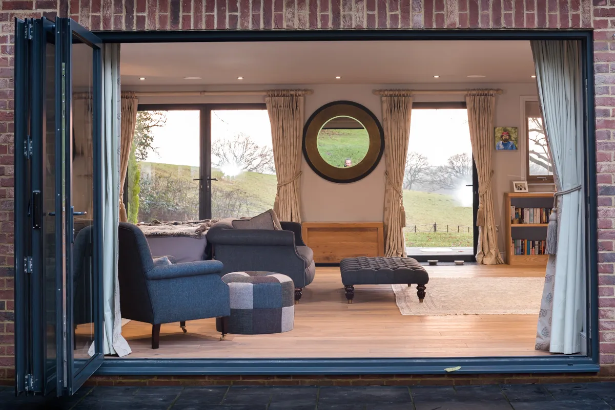 fully open bifold door from the outside looking in to a room with sofas, foot stools, wood flooring and glass windows and door on the opposite wall