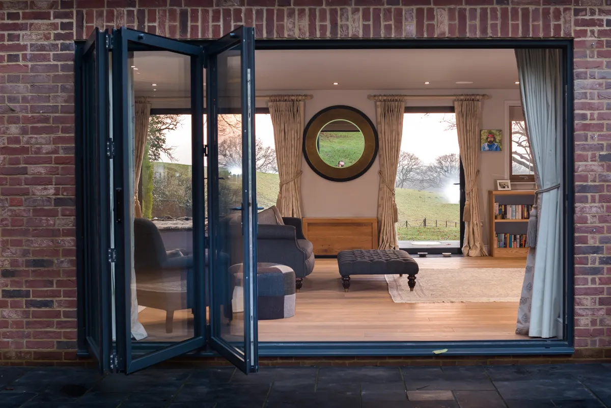 Slighlty closed bifold door from the outside looking in to a room with sofas, foot stools, wood flooring and glass windows and door on the opposite wall