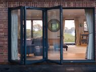 half closed bifold door from the outside looking in to a room with sofas, foot stools, wood flooring and glass windows and door on the opposite wall