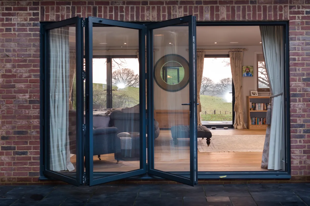 half closed bifold door from the outside looking in to a room with sofas, foot stools, wood flooring and glass windows and door on the opposite wall