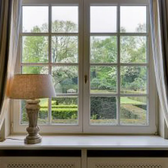 a upvc window viewed from inside looking out onto mature gardens.  window is framed with curtain.  a lit lamp sits to the left side of a deep window shelf-