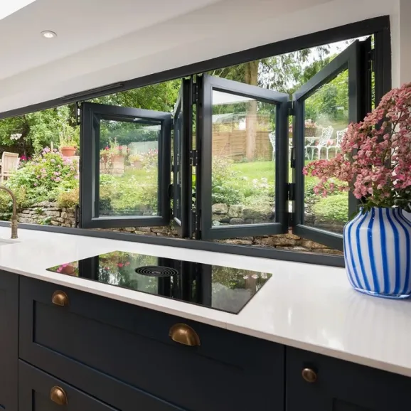 partially opened bifold window viewed from inside a kitchen at the counter looking out the window into a garden. A blue and white striped vase with pink fl